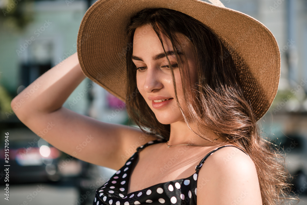 Pretty girl in a black dress and a hat with wide flaps smiles enjoying walk down the European town during sunny summer day. Traveling in warm season.