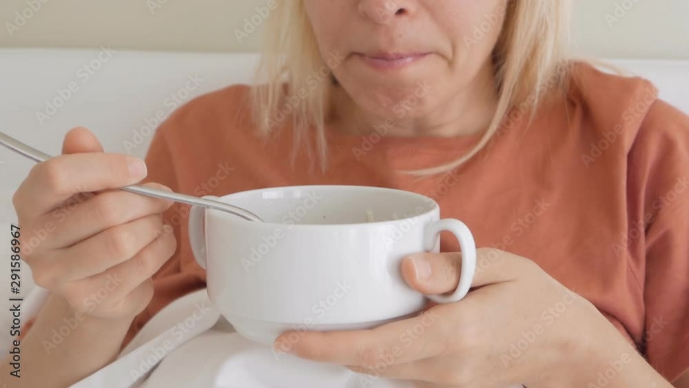 A woman with a sad painful face sitting on bed under white blanket and eating from a plate of soup.