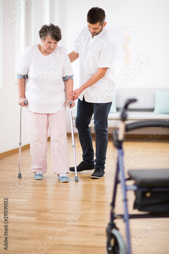 Senior woman at hospital learning how to walk on crunches and supporting male nurse © Photographee.eu