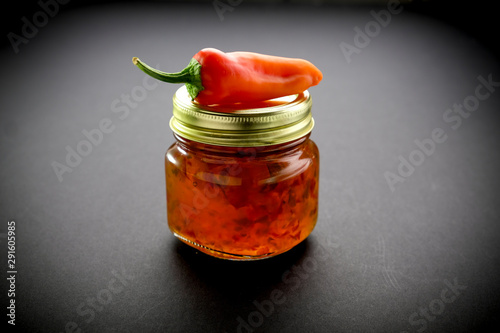 Small glass jar of jalapeño jelly illuminated by light from rear and side with a red banana pepper on top of jar photo