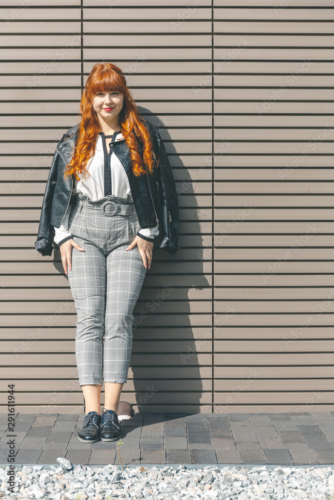 Cute young girl in a black leather jacket with curly red hair and fringe stands near a modern wall on a sunny day and looks at the camera