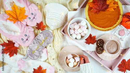 Cozy autumn Thanksgiving in bed flatlay overhead with cosy sweater, slippers, socks and tray with pumpkin pie and hot chocolate with marshmallows.