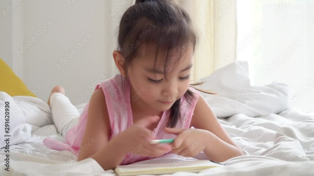 4K Medium shot happy smiling little cute asian preschool child girl lying down on the bed enjoy doing homework or writing on the book. Relaxing learning homeschool education and back to school concept 4K Medium shot happy smiling little cute asian preschool child girl lying down on the bed enjoy doing homework or writing on the book. Relaxing learning homeschool education and back to school concept