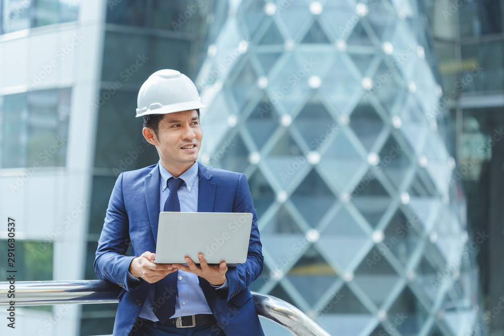 Asian engineer with white hard hat holding laptop working outdoor place.