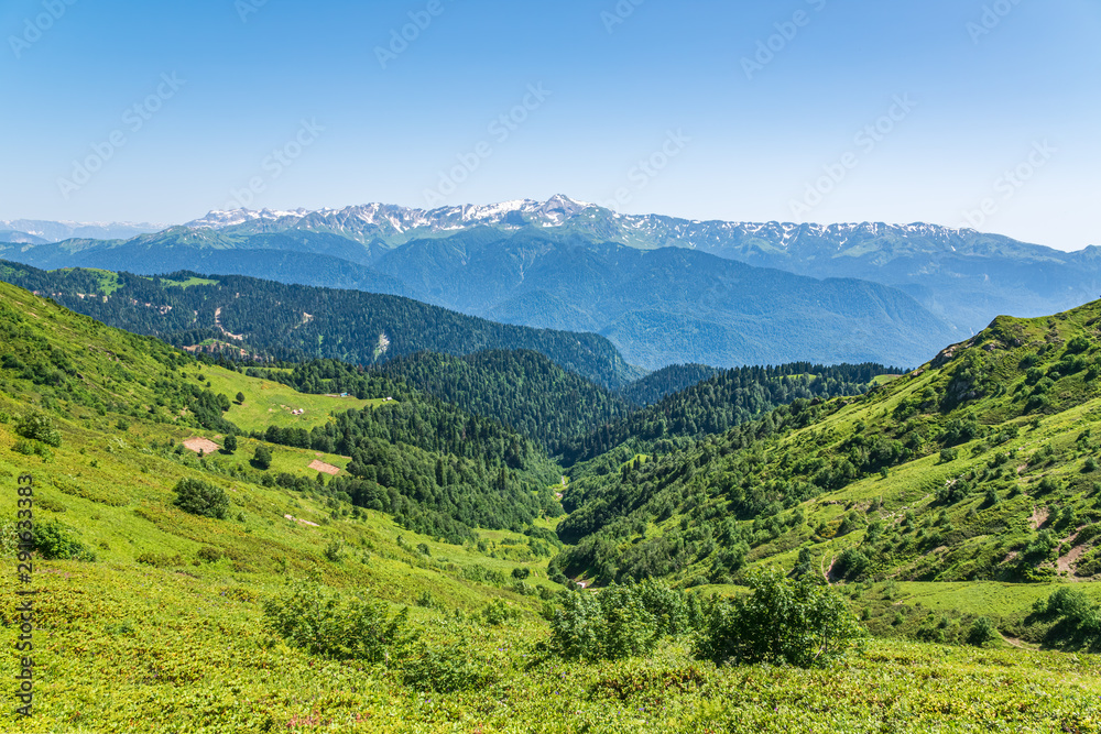 The view from the height to the green mountain valley surrounded by high mountains. Snow-capped mountain peaks on the horizon. Krasnaya Polyana, Sochi, Caucasus, Russia.