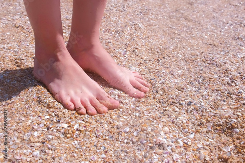 Barefooted woman massaging her feet on sandy beach in seashells at seaside. Nature massage for legs on seashore. Girl traveller on sea vacation. Tourist rest relax concept. Wellness treatment on cost.