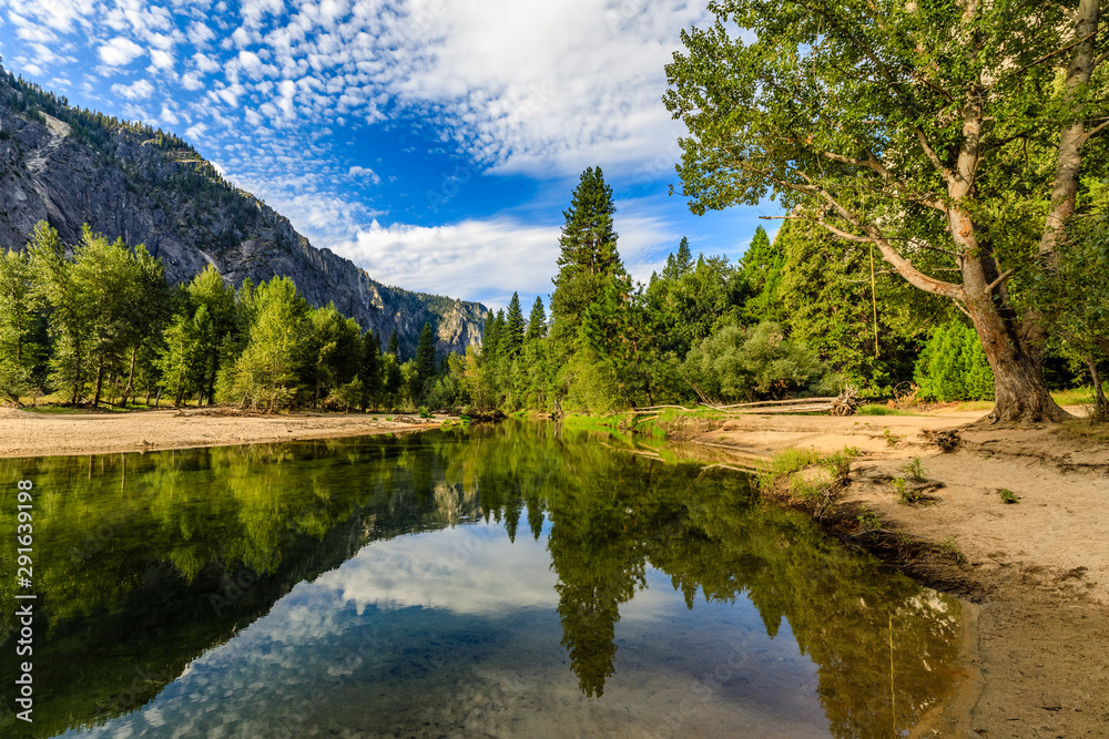 Sandy beaches and lush green trees reflect from both sides of the Merced River in Yosemite national park