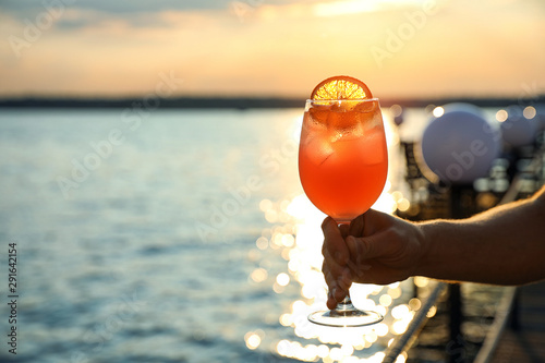 Man with glass of fresh summer cocktail near water at sunset, closeup. Space for text
