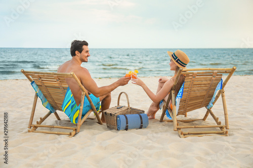 Happy young couple with cocktails sitting on deck chairs at sea beach