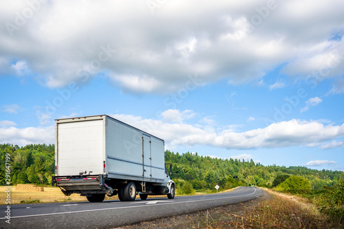 Big rig semi truck with long box trailer driving on the winding road with forest and meadow