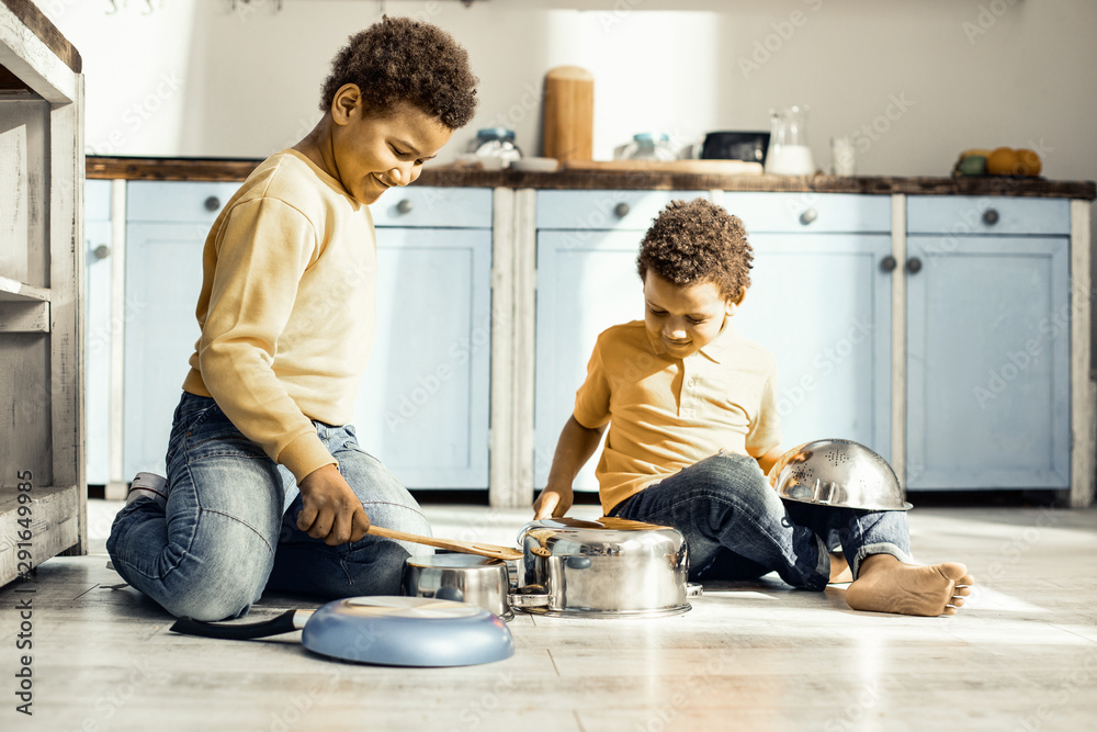 Fototapeta premium Two boys sitting on the floor of the kitchen and playing with the pans.