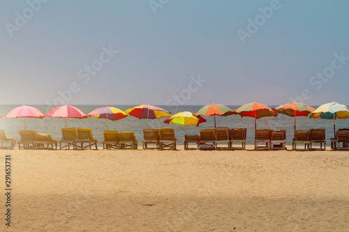 Row of covered wooden deck chairs with colorful umbrellas on the beach in GOA  India  toned. Sea summer template. Long shot.