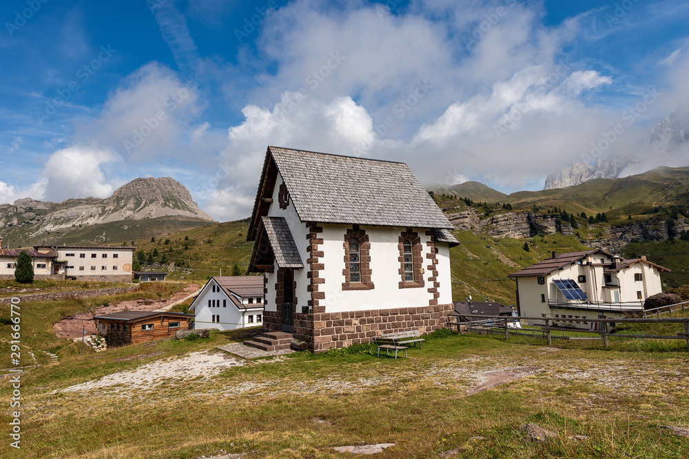 Fototapeta premium Small church with the Pale di San Martino and Mount Castellaz in the background. Passo Rolle, Fiera di Primiero, Dolomites, Italian Alps, Trentino Alto Adige, Italy, Europe