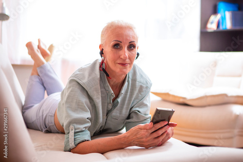 Adults and Technologies Concepts. Positive Caucasian Senior Woman Posing with Smartphone and Headphones On Couch Indoors.