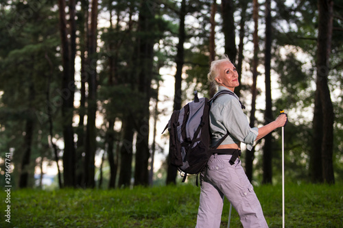 Senior Sports and Healthy Lifestyle Concepts. Portrait of Positive Mature Caucasian Woman Having Fitness Nordic Walking Exercise with Backpack and Sticks in Forest.