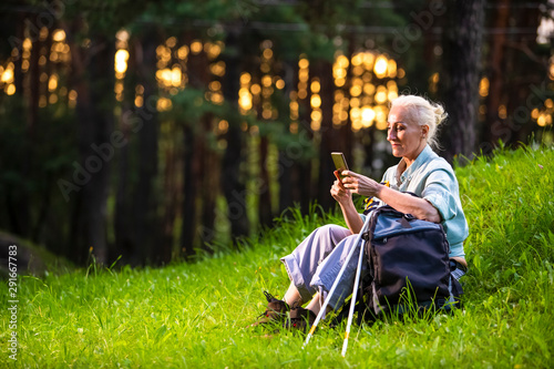 Senior Healthy Lifestyle Concepts. Portrait of Positive Mature Caucasian Woman Having Fitness Nordic Walking Exercise with Backpack and Sticks in Forest. Working with Smartphone. photo