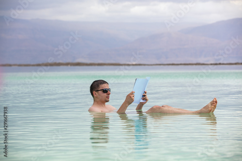 Man swimming in the sea and reading a magazine during spa procedures. Summertime, holiday, travel concept. Dead Sea scene