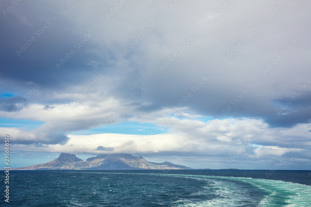 Island on the horizon. Rocks in the sea. Beautiful rocky sea landscape with dramatic cloudy sky. Water trail foaming behind a ferry. View from the ship. Nature of Norway