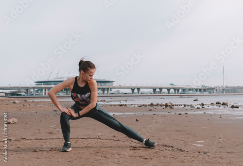 young slender woman in sportswear doing stretching on the beach in the background of the city, training, warmup at the seaside