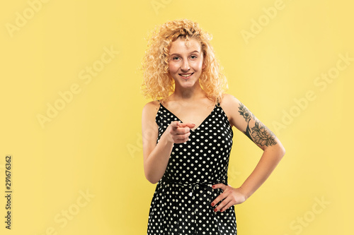 Caucasian young woman's half-length portrait on yellow studio background. Beautiful female model in black dress. Concept of human emotions, facial expression. Showing and pointing something. Inviting.