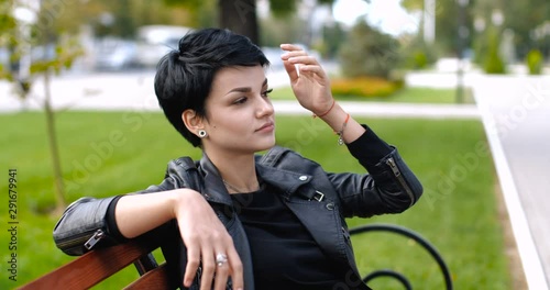 Attractive Caucasian short-haired brunette girl sitting on a bench in a city park, straightening her hair and smiling looking around. 4K slow motion 50 fps slide camera