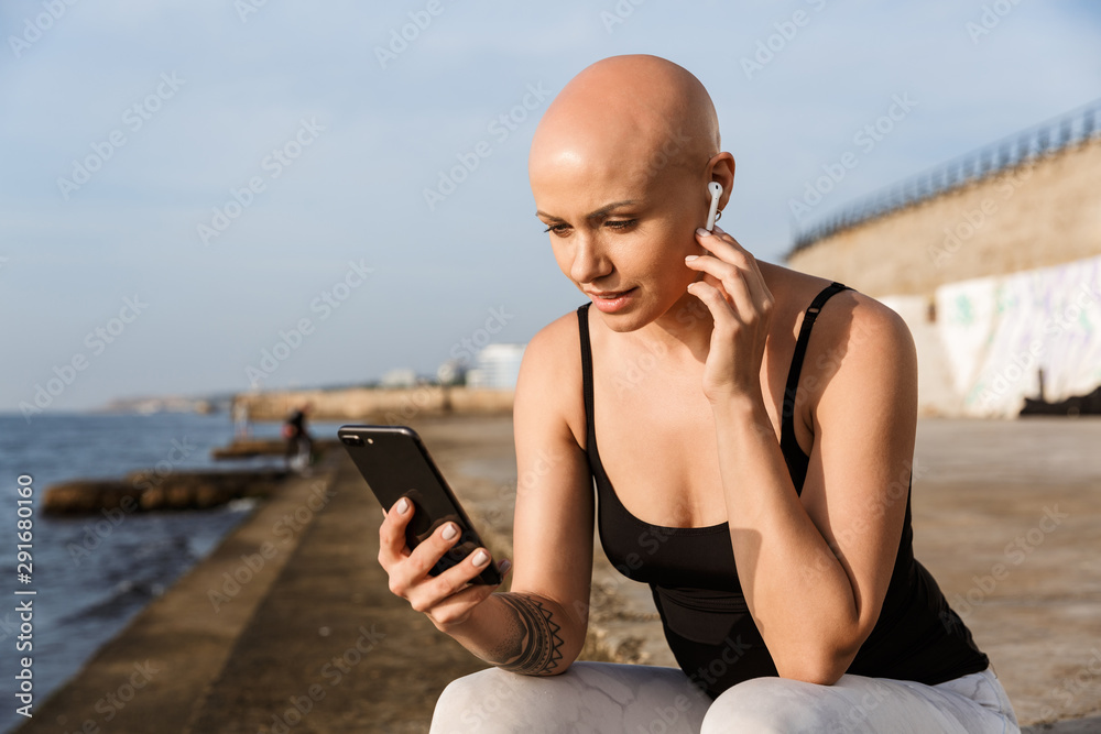 Image of pleased bald woman using cellphone and earphone while sitting