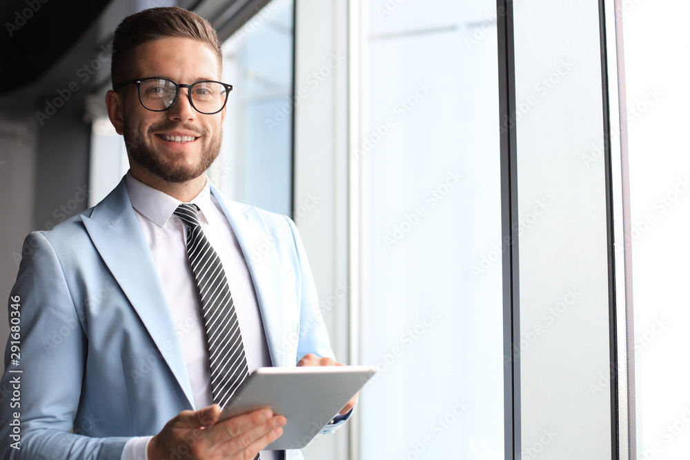 Modern business man in formalwear using digital tablet while standing near window in the office Modern business man in formalwear using digital tablet while standing near window in the office