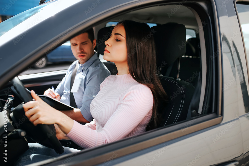 Male auto instructor takes exam in young woman. She hold hands on steering wheel and driving. Young man look at brunette with confusion. Model drives straight. Sit in car.