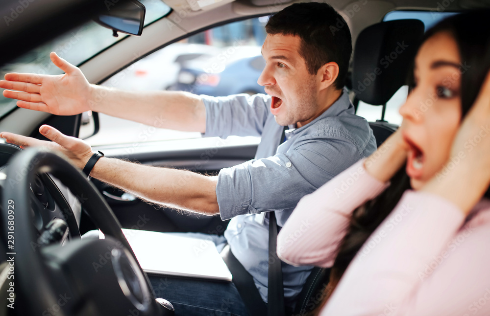 Male auto instructor takes exam in young woman. Stress during driving. Scared young woman touch head with hands. Instructor screaming and pointing forward.