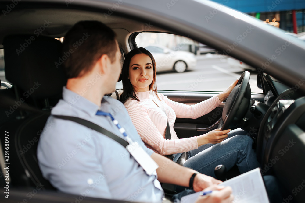 Male auto instructor takes exam in young woman. Calm peaceful confident female student look at teacher and smile. Hold hands on steering wheel and drive. Male auto instructor takes exam in young woman. Calm peaceful confident female student look at teacher and smile. Hold hands on steering wheel and drive.
