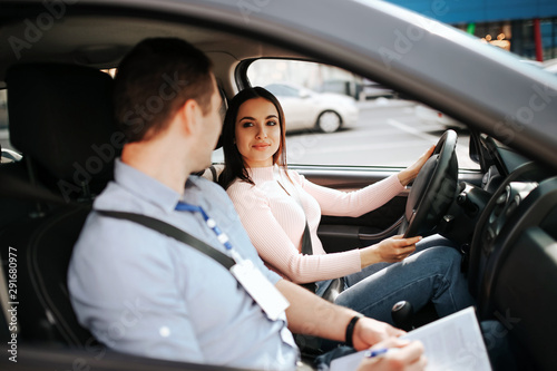 Male auto instructor takes exam in young woman. Calm peaceful confident female student look at teacher and smile. Hold hands on steering wheel and drive.
