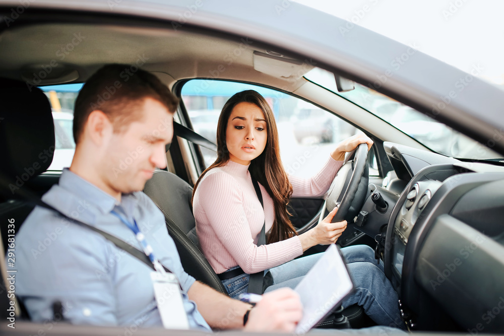 Male auto instructor takes exam in young woman. Serious concentrated guy writing on paper. Wondered woman look at it. Holding hands on steering wheel. Passing practical test.