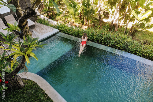 Fototapeta Naklejka Na Ścianę i Meble -  Back view of woman in red bikini relaxing in infinity pool in Bali admires a beautiful view of the palm trees.Luxury holiday.Girl resting on the island of Bali. Copy space. Vacation concept
