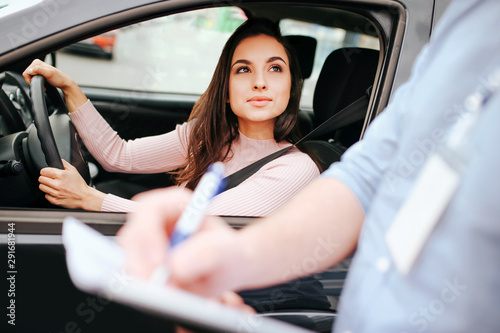 Male auto instructor takes exam in young woman. Guy writes test result in paper. Interesting woman look at guy outside from car. Hold hands on steering wheel and seat belt across body.
