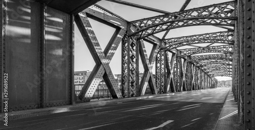 Fototapeta Naklejka Na Ścianę i Meble -  Black and white asphalt road under the steel construction of a bridge in the city on a sunny day. Evening urban scene with the sunbeam in the tunnel. City life, transport and traffic concept.