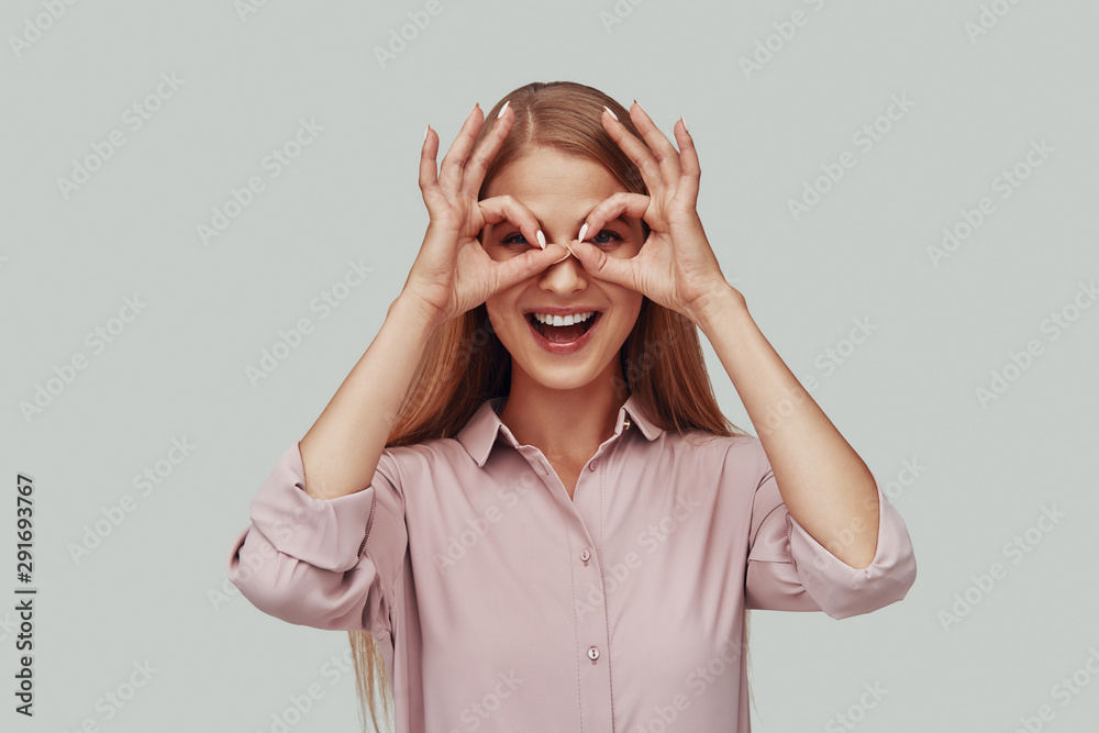 Playful young woman looking at camera and smiling while standing against grey background Playful young woman looking at camera and smiling while standing against grey background