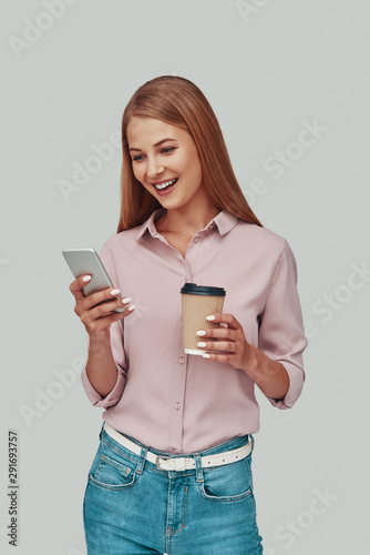Attractive young woman using smart phone and smiling while standing against grey background