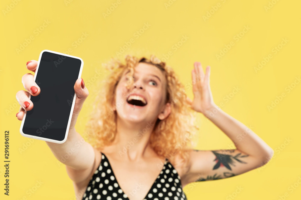 Naklejka premium Caucasian young woman's half-length portrait on yellow studio background. Beautiful female model in black dress. Concept of human emotions, facial expression. Showing phone's screen, looks crazy happy