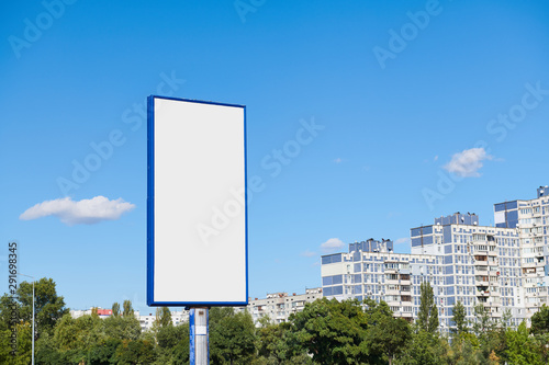Big vertical billboard with copy space on urban background against blue sky. Advertising space with high-rise building and trees.