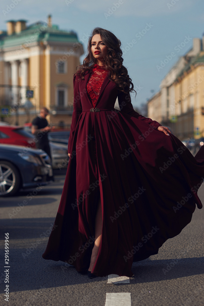 Naklejka premium Young beautiful stunning woman in red luxury evening dress with flying hem walking city street on a sunny evening. Elegant lady with makeup and wavy brunette hair. Full length fashion portrait