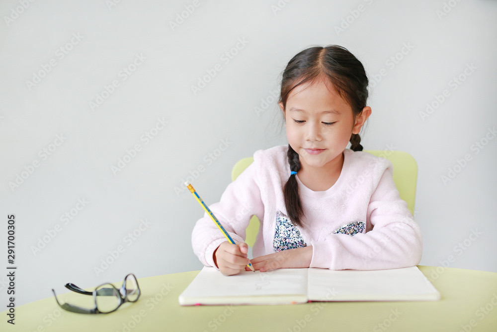 Happy little Asian child girl write in a book or notebook with pencil sitting on kid chair and table against white background. Happy little Asian child girl write in a book or notebook with pencil sitting on kid chair and table against white background.