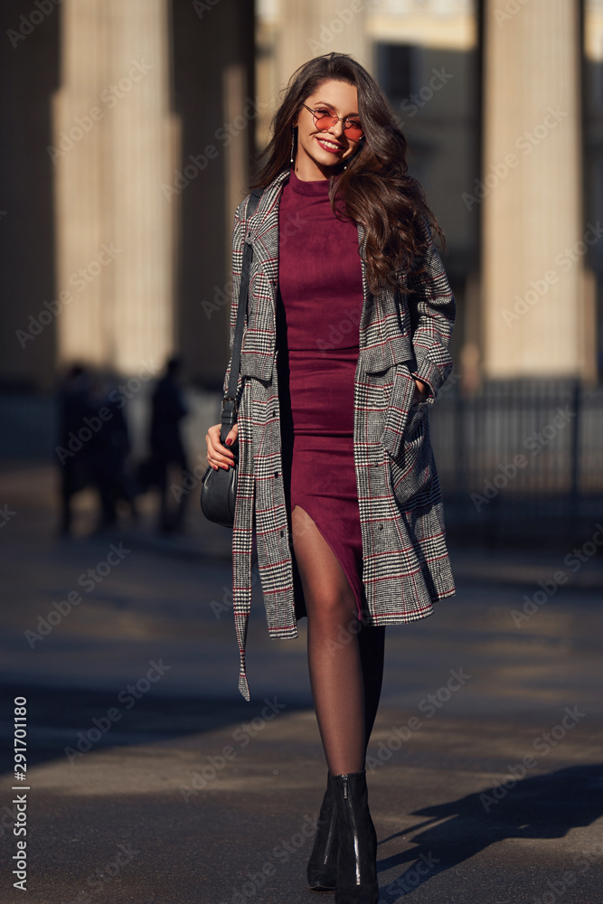 Full length outdoot portrait of young elegant stylish woman in burgundy color dress, gray coat and sunglasses walking in city on a sunny day. Elegant girl with dark wavy hair.