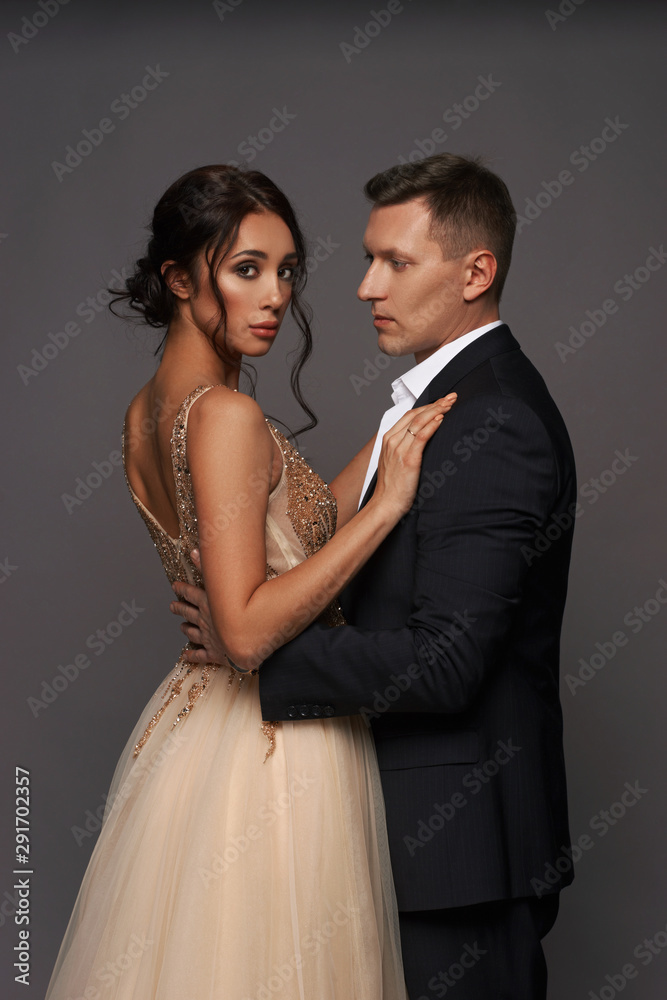 Attractive beautiful and well-dressed young adult couple posing in studio on gray background. Woman in beautiful evening dress and man wearing black classical suit with white jacket
