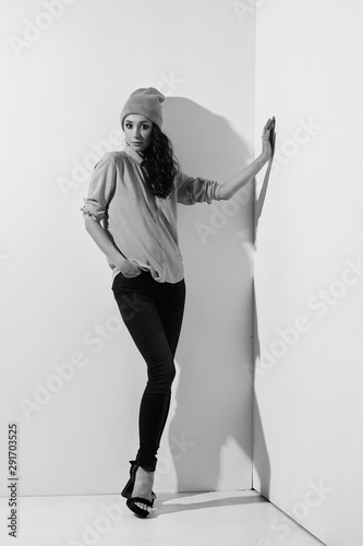 Casual dressed woman standing in white corner. Brunette girl with long vawy hair posing in black jeans, yellow blouse and yellow hat. photo