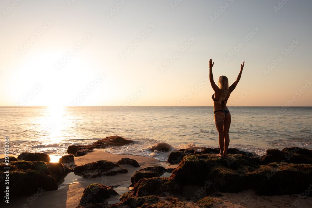 Fototapeta premium Excited young woman raising arms at the beach in front of the ocean. View from back. Sunset at the beach. Bali, Indonesia.