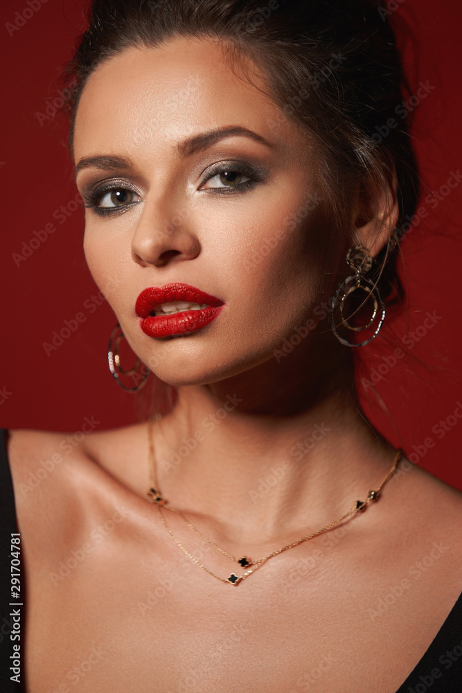Closeup fashion vogue style portrait of young elegant woman with hairstyle and makeup. Girl with red lips wearing earrings and necklace. Studio shot against red background Closeup fashion vogue style portrait of young elegant woman with hairstyle and makeup. Girl with red lips wearing earrings and necklace. Studio shot against red background