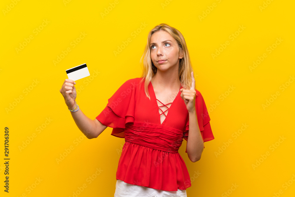 Blonde young woman over isolated yellow background holding a credit card Blonde young woman over isolated yellow background holding a credit card
