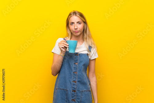 Blonde young woman over isolated yellow background holding hot cup of coffee