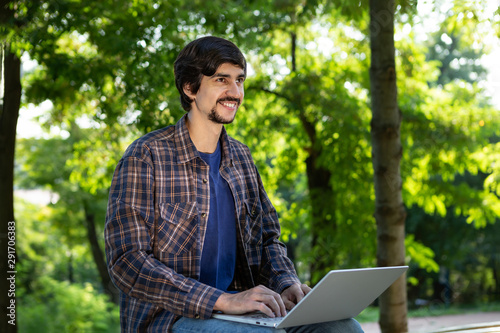 Young bearded brunet freelancer with mustache sitting with a laptop in a park. Work anywhere