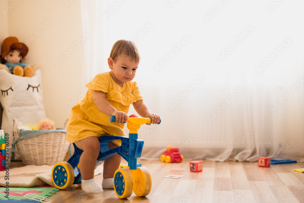 Cute lovely small baby girl learning to balance on her first bike at home. Toddler activity at daycare - obrazy, fototapety, plakaty 